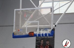 Canasta de baloncesto en una cancha cubierta, sin jugadores en la imagen, con estructura metálica y red de protección en el fondo.