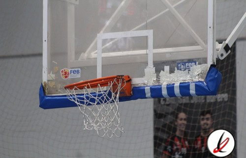 Balón de baloncesto en el aro en una cancha cubierta, con espectadores en el fondo.