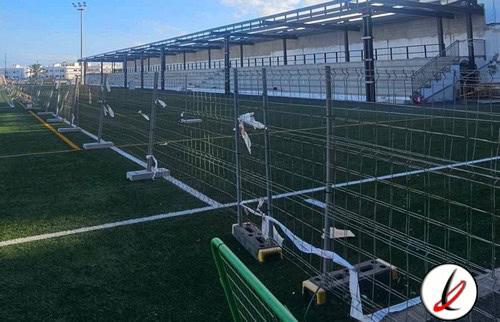 Barandilla de protección en un campo de fútbol con gradas y estructura metálica en el fondo, en un estadio al aire libre.