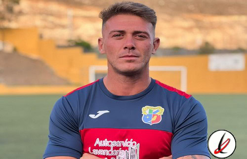 Jugador de fútbol con camiseta azul y roja en un campo de césped, con fondo de gradas y muro en Lanzarote.
