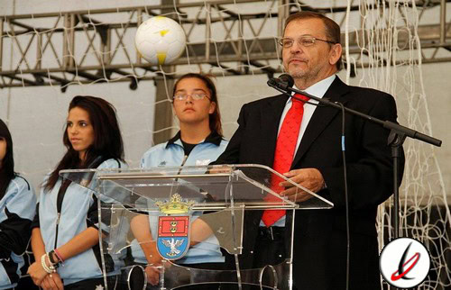 Hombre en traje y corbata hablando en un acto oficial con dos jóvenes en un escenario con red de voleibol y balón, en un evento público en Lanzarote.
