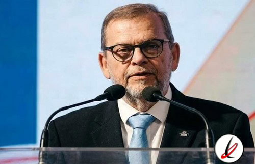 El hombre con gafas y barba, vestido con traje y corbata, hablando en un micrófono en un evento formal.
