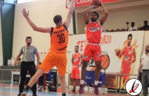 Jugador con camiseta roja saltando para encestar en un partido de baloncesto, mientras un defensor con camiseta naranja intenta bloquear el tiro.