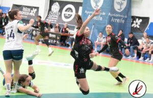 Jugadoras de voleibol en acción durante un partido en un pabellón deportivo, con público y banners publicitarios en el fondo.