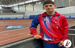 Atleta joven en pista de atletismo cubierta, con ropa deportiva roja y azul, en un estadio con pista de carreras y anuncios publicitarios en el fondo.