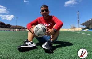 Hombre sonriendo con gafas y camiseta roja, sentado en un campo de fútbol con balón, en un estadio al aire libre bajo cielo azul.