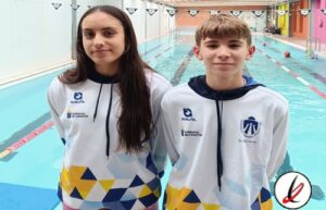 Dos jóvenes nadadores con camisetas de equipo en una piscina cubierta, listos para competir o entrenar, con fondo de carriles y paredes de colores en el centro acuático.