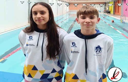 Dos jóvenes nadadores con camisetas de equipo en una piscina cubierta, listos para competir o entrenar, con fondo de carriles y paredes de colores en el centro acuático.