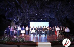 Personas en un escenario en una ceremonia de premiación en una cueva, con un grupo de individuos sosteniendo certificados y trofeos, en un evento formal y cultural.