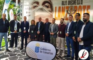 Grupo de personas en un evento deportivo en Lanzarote, con un cartel del Gobierno de Canarias y un fondo con una imagen de un rostro y texto promocional.