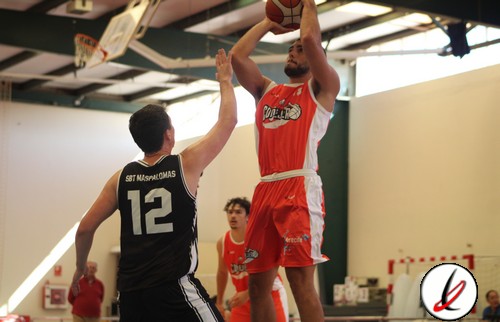 Jugador de baloncesto en acción lanzando el balón en una cancha cubierta, con defensor intentando bloquear el tiro, en un partido de baloncesto en interiores.