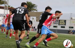 Jugadores de fútbol en acción durante un partido en un campo al aire libre, con un fondo de gradas y árboles, en un día soleado y despejado.