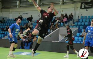 Saltador en acción durante un partido de balonmano en un pabellón cubierto, con jugadores en el suelo y espectadores en las gradas.