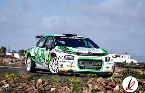 Línea 1: Coche de rally con patrocinadores en un terreno rocoso, bajo cielo nublado.