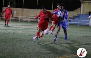 Jugadores de fútbol en acción durante un partido en un campo al aire libre, con espectadores y gradas en el fondo, en un entorno nocturno.