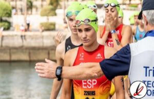 Nadadoras en la playa preparándose para la competencia, con un entrenador dando instrucciones, todas llevan gorros de natación verdes y trajes rojos, en un entorno al aire libre junto al agua.