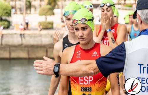 Nadadoras en la playa preparándose para la competencia, con un entrenador dando instrucciones, todas llevan gorros de natación verdes y trajes rojos, en un entorno al aire libre junto al agua.