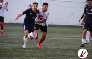 Jugadores de fútbol en acción durante un partido en un campo de césped artificial, con fondo de muro blanco y portería.