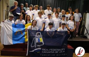 Grupo de niños y jóvenes deportistas con banderas en un escenario, acompañados por adultos, en un evento deportivo en Lanzarote.