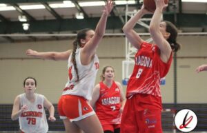 Taga de baloncesto femenina en acción durante un partido en un gimnasio cubierto. Jugadoras en uniforme rojo y blanco compiten por la posesión del balón, con una jugadora en el aire intentando encesta.