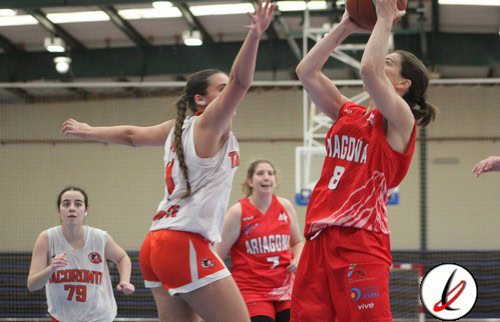 Taga de baloncesto femenina en acción durante un partido en un gimnasio cubierto. Jugadoras en uniforme rojo y blanco compiten por la posesión del balón, con una jugadora en el aire intentando encesta.